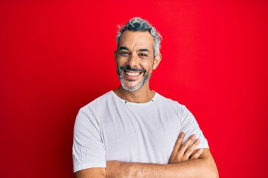 Orta yaşlı, beyaz tişört giyen, kollarını kavuşturup kameraya gülümseyen gri saçlı bir adam. pozitif kişi. 