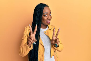 Sıradan bir ceket giyen, gülümseyen, parmaklarını zafer işareti yaparken gösteren genç Afrikalı Amerikalı kadın. İki numara.. 