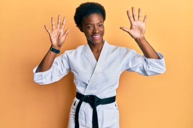 Genç Afrikalı Amerikalı kız karate kimonosu ve siyah kuşak takıyor ve parmaklarını on numarayı gösterirken kendinden emin ve mutlu bir şekilde gülümsüyor.. 