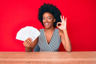 Genç Afro-Amerikalı kadın elinde dolarlarla tamam işareti yapıyor, gülümsüyor ve mükemmel bir sembol oluşturuyor. 