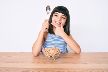 Bangır bangır küçük bir kız masada oturmuş mısır gevreği yiyor. Gülümsüyor, eli ve parmağı ile işaret ediyor. 