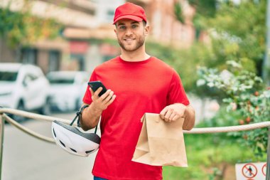 Beyaz atlı genç teslimatçı akıllı telefon kullanıyor ve şehirde kağıt torba taşıyor..
