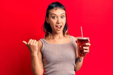 Bir bardak kola içen genç bir kadın başparmağını yana kaldırarak mutlu bir şekilde gülümsüyor. 