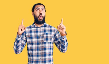 Gündelik giysiler giyen genç Arap şaşırmış ve şaşırmış bir şekilde yukarı bakıyor ve parmaklarıyla işaret ediyor ve kollarını kaldırıyordu.. 