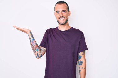 Dövmeli, sıradan kıyafetler giyen, neşeli gülümseyen, eli avucunda kameraya bakan genç ve yakışıklı bir adam.. 