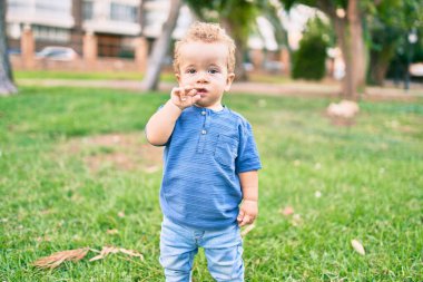 Üzgün küçük çocuk, güneşli bir günde parkta diş etlerine dokunduğu için parmaklarını dudaklarına koyuyor. Güzel sarı saçlı erkek bebek yeni bebek dişleri için acı çekiyor.