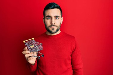 Genç İspanyol adam elinde küçük bir süpermarket alışveriş arabası tutarken düşünceli ve ayık bir ifade takınıyor. Kendinden emin görünüyor. 