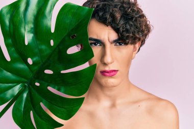 Güzel yüzüne yakın yeşil bitki yaprağı tutan, makyaj yapan genç bir adam şüpheci ve gergin, sorun yüzünden somurtuyor. negatif kişi. 