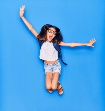 Sevimli İspanyol öğrenci kız gözlüklü ve ağzı açık sırt çantalı. Kolları açık atlama heyecanı izole edilmiş mavi arka planda