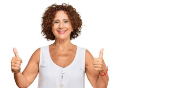 Güzel, orta yaşlı olgun bir kadın, günlük beyaz gömlek giyer, başarı işareti yapar, elleriyle olumlu jest yapar, gülümser ve mutlu olur. Neşeli ifade ve kazanan jest. 