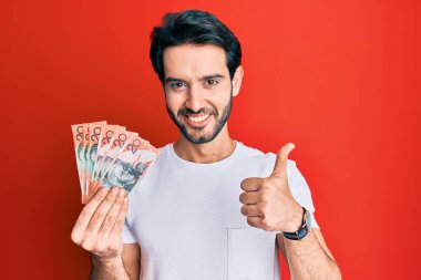 Avustralya doları tutan İspanyol asıllı genç adam mutlu ve pozitif gülümsüyor, başparmağıyla mükemmel ve onay işareti yapıyor. 