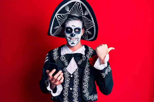 年轻人穿着墨西哥式的死亡服装 用智能手机把大拇指指向旁边 高兴地张开嘴 — 图库照片