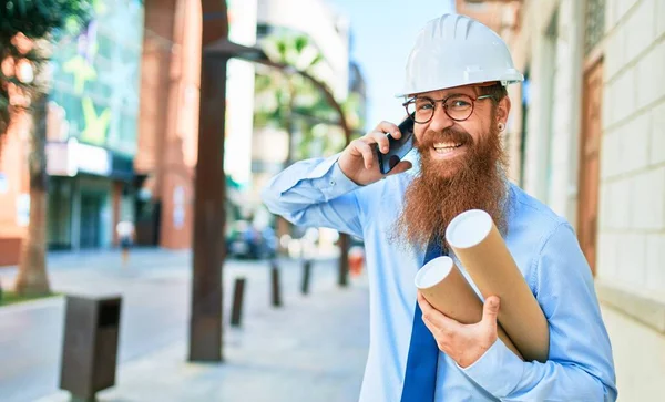 長いひげを生やした若い赤毛の建築家の男は ハード帽子を笑顔幸せを着ている 街の通りで設計図を持っているスマートフォンで会話をしていると — ストック写真