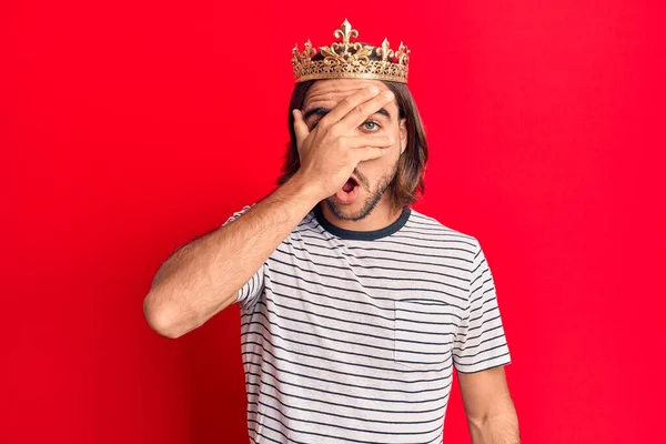 Young handsome man wearing prince crown peeking in shock covering face and eyes with hand, looking through fingers afraid