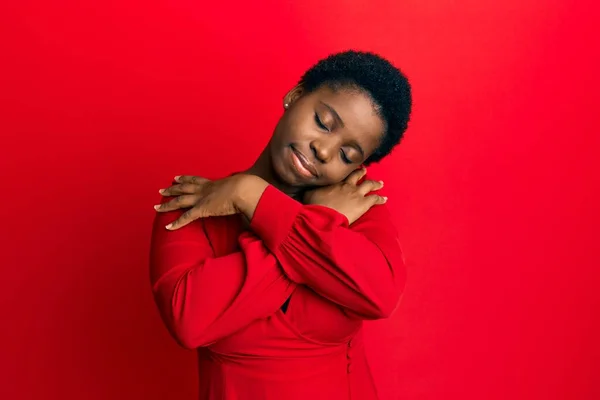 年轻的非洲女人 一头黑发 穿着休闲的衣服 披着红色的背景 幸福而积极地拥抱自己 充满自信地微笑 自爱和自我照顾 — 图库照片