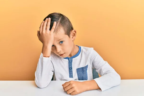 Adorable caucasian kid wearing casual clothes sitting on the table surprised with hand on head for mistake, remember error. forgot, bad memory concept.