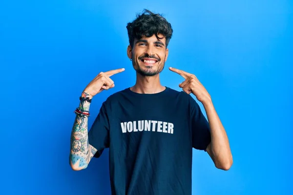 ボランティアのTシャツを着た若いヒスパニック系の男性は 陽気な笑みを浮かべて指の歯と口を指しています 歯の健康の概念 — ストック写真
