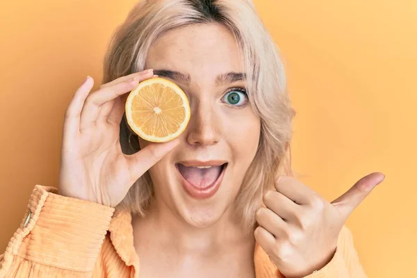 Young Blonde Girl Holding Lemon Eye Pointing Thumb Side Smiling — ストック写真