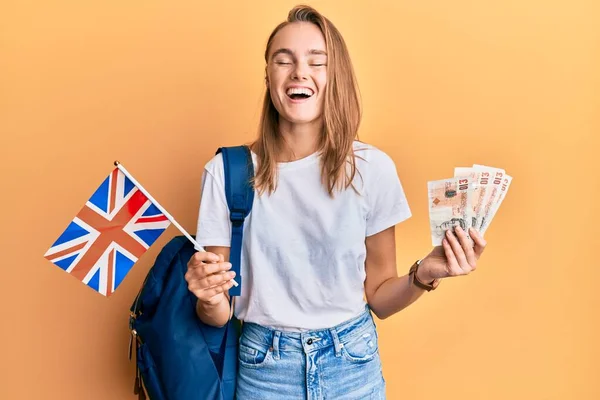 Beautiful blonde woman exchange student holding uk flag and pounds smiling and laughing hard out loud because funny crazy joke.