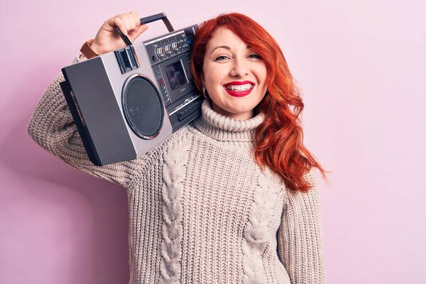 Beautiful redhead woman listening to music holding vintage boombox over pink background looking positive and happy standing and smiling with a confident smile showing teeth