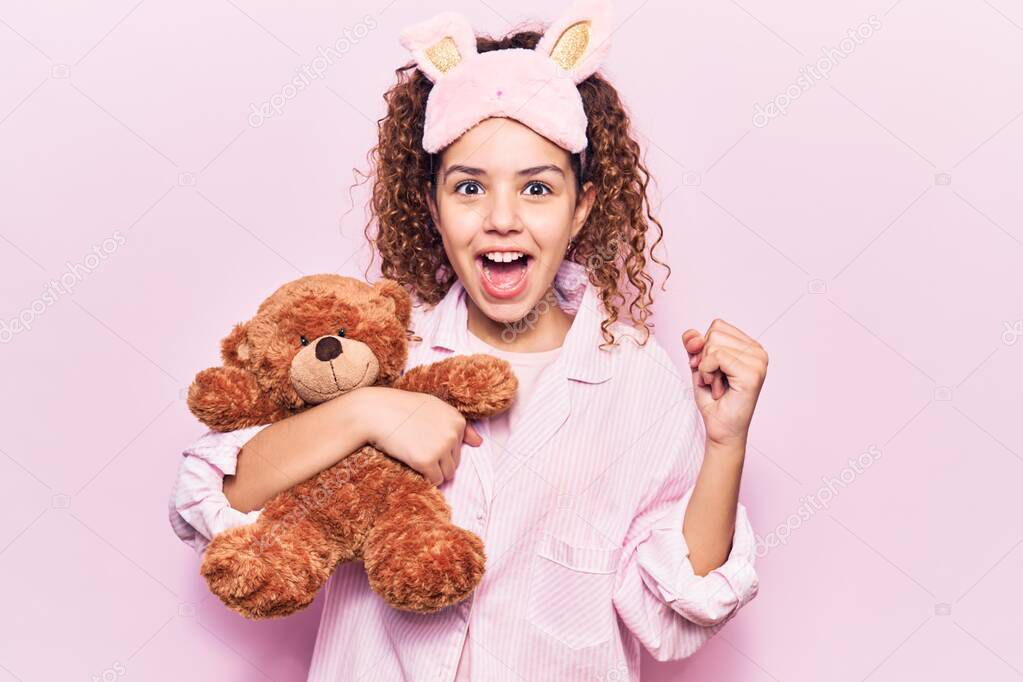 Beautiful kid girl with curly hair wearing sleep mask and pajamas holding teddy bear screaming proud, celebrating victory and success very excited with raised arms 