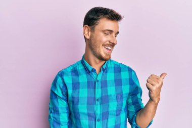 Yakışıklı beyaz adam, günlük kıyafetler giyiyor, gülümsüyor, gülümsüyor ve başparmağıyla yan tarafı işaret ediyor.. 