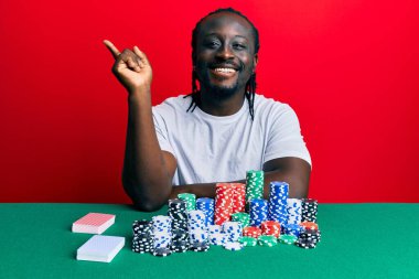 Yakışıklı genç siyah adam poker fişleri ve kartlarla masada oturuyor mutlu mesut gülümsüyor. 