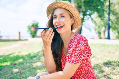 Genç sarışın kadın tatilde gülümsüyor. Parkta oturmuş akıllı telefondan konuşuyor..