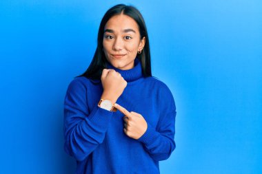 Genç Asyalı kadın, günlük kışlık süveter giyiyor. Aceleyle saati, sabırsızlığı ve rahat bir ifadeyle kameraya bakıyor. 