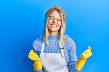 Güzel sarışın kadın daha temiz önlük ve eldiven giyer. Çok mutlu ve heyecanlı. Kolları havada, gülümseyen ve başarı için çığlık atan kazanan bir jest yapıyor. kutlama konsepti. 