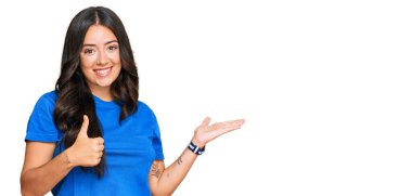 Güzel, esmer, günlük kıyafetler giyen, avuç içi gösteren ve baş parmaklarıyla hareket eden mutlu ve neşeli gülümseyen genç bir kadın. 