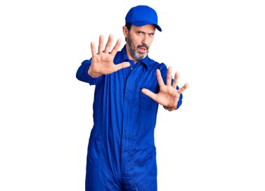 Orta yaşlı, tamirci üniforması giyen yakışıklı bir adam avuç içi hareketlerini durduruyor, kızgın ve hayal kırıklığına uğramış bir ifade takınıyor. 