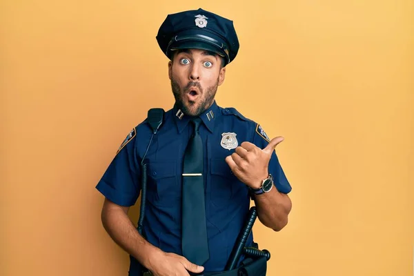 Handsome hispanic man wearing police uniform surprised pointing with hand finger to the side, open mouth amazed expression.