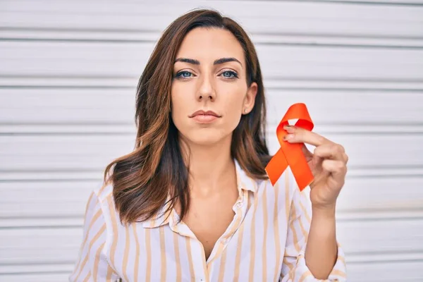 Young hispanic woman with serious expression holding awareness orange ribbon standing at the city.