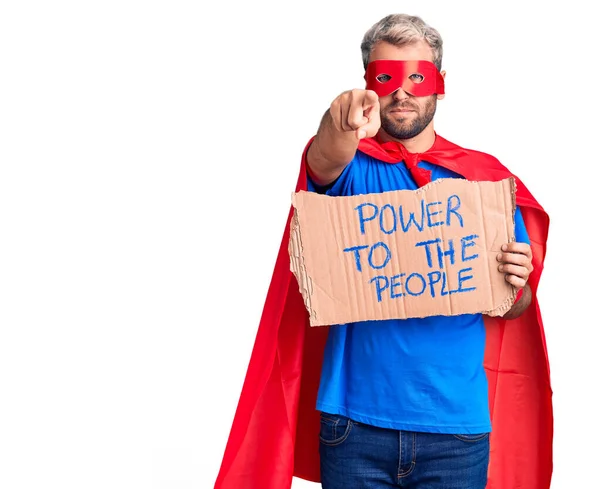 Young blond man wearing super hero custome holding power to the people cardboard banner pointing with finger to the camera and to you, confident gesture looking serious