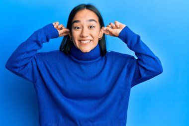 Kış süveteri giyen genç Asyalı kadın gülümsüyor kulakları parmaklarıyla çekiyor, komik bir jest. deneme sorunu 