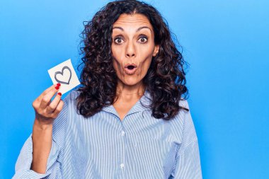 Orta yaşlı güzel bir kadın, elinde kalp anımsatıcı, korkmuş ve şaşırmış bir şekilde ağzı açık, şaşırmış, inançsız bir yüz ifadesi var. 