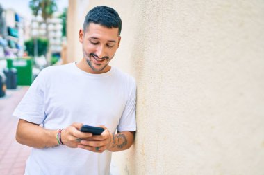 Duvara dayanmış akıllı telefon kullanan, mutlu gülümseyen genç İspanyol adam..