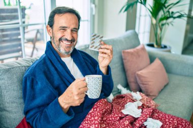 Orta yaşlı bir adam evde üşütmüş ve ateşi var. Kanepede oturan grip hastası.