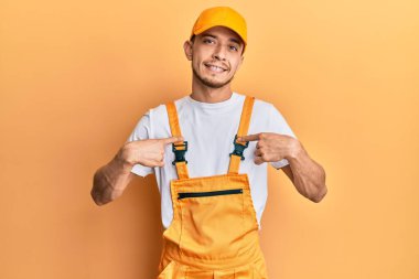 Tamirci üniforması giyen İspanyol genç adam. Yüzünde bir gülümsemeyle kendinden emin görünüyor. Kendini gururlu ve mutlu olarak gösteriyor.. 