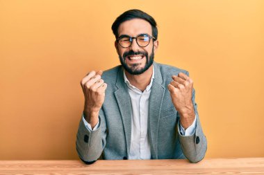 Ofiste çalışan çok mutlu ve heyecanlı genç İspanyol adam kollarını kaldırıp, gülümseyerek ve başarı için bağırarak kazanan jestler yapıyor. kutlama konsepti. 