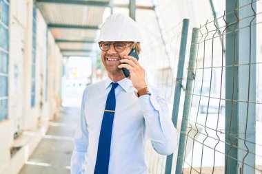 Bir inşaat projesinin önünde dikilirken kasketli mimar telefonda konuşuyor.