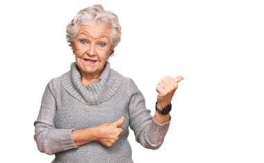 Kış süveteri giyen yaşlı, gri saçlı kadın arka tarafı işaret ediyor. El ve baş parmakları havada, kendinden emin gülümsüyor. 