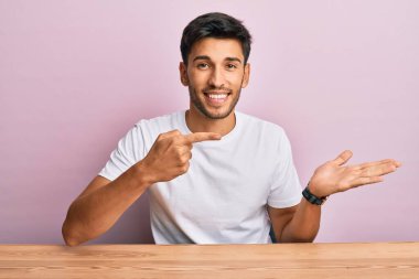 Masada oturan, sıradan beyaz t-shirt giyen yakışıklı genç adam hayrete düşmüş ve kameraya gülümsüyordu. Elle ve parmakla gösteriyordu.. 