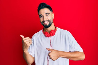 Sakallı, müzik dinleyen, kulaklık takan, arka tarafı işaret eden, el ve baş parmakları havada, kendinden emin genç bir adam. 