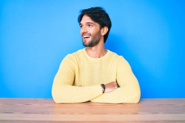 Yakışıklı İspanyol bir adam, sıradan bir süveter giyer, masaya oturur, yüzünde gülümsemeyle yan yana bakar, doğal bir ifade. Kendinden emin gülen. 