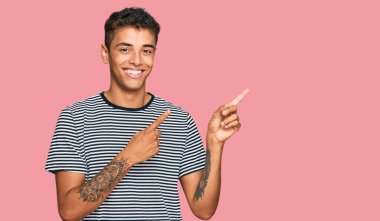 Genç, yakışıklı, Afro-Amerikalı sıradan kıyafetler giyen, gülümseyen ve kameraya bakıp iki elini ve parmağını yana doğru gösteren bir adam.. 