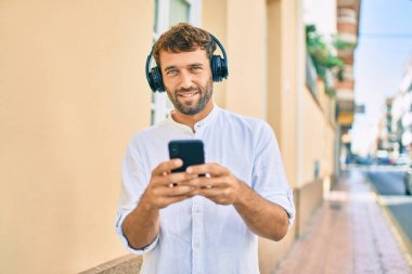 Güneşli bir günde sakallı yakışıklı bir adam açık havada mutlu bir şekilde gülümsüyor kulaklık takarak müzik dinliyor.