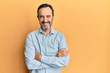 Orta yaşlı İspanyol bir adam sıradan kıyafetler giyip kollarını kavuşturup kameraya bakarken gülümsüyor. pozitif kişi. 