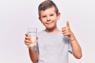 Sevimli sarışın çocuk bir bardak su içiyor mutlu ve pozitif gülümsüyor, başparmağı mükemmel ve onay işareti yapıyor. 